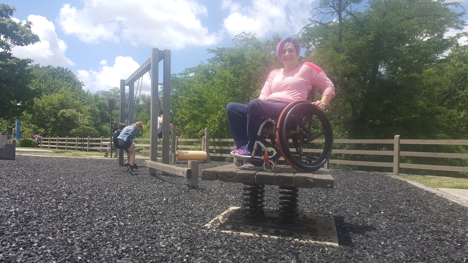 Allaina seated in her wheelchair on an accessible elevated play platform while children play nearby